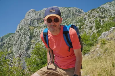 Man in an coral hiking shirt and cap resting on a steep mountain trail in Montenegro, with rocky peaks and summer vegetation in the background